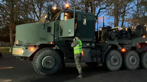 A member of military personnel in a hi-vis jacket, stood next to the cabin of a very large lorry