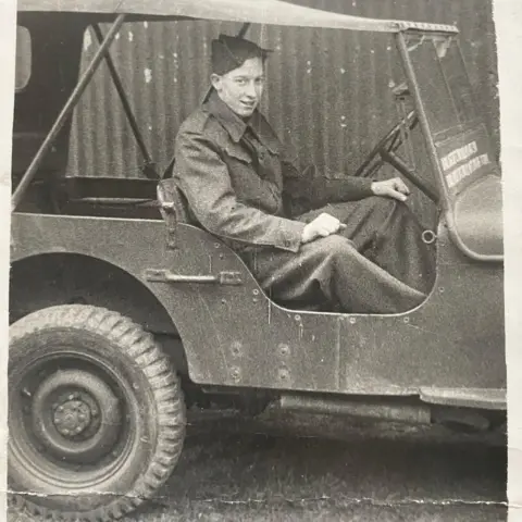 An old black and white photo of a young man in army uniform driving an old army jeep.