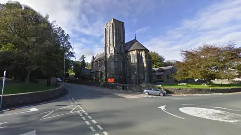 A flat roundabout to the right sits between three roads. Saddle Road runs from right to left, with a large stone church to the righthand side of the walled roads, with trees on the left. It is a sunny day with some whispy clouds.