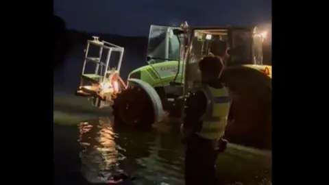 Hannah Waddington A police officer standing on the shore of Windermere while a tractor enters the water. It is dark outside.