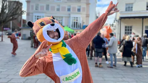 Simon smiles at the camera as he wears a full body squirrel costume and he's doing Usain Bolt's winning pose. Simon has a yellow and green scarf on and people are gathered behind him and some are also dressed as squirrels.