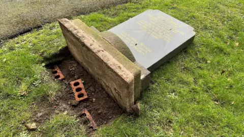 A grave headstone uprooted from the ground. Bricks are visible beneath the stone.