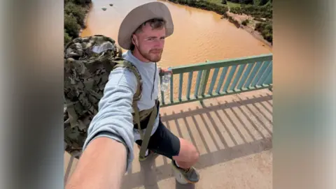 James Lewis Lewis is standing on a bridge over a muddy-looking river. He is in shorts, a top and a sunhat. He has a short beard and is taking the selfie from a tilted angle.