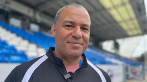 Emma Baugh/BBC Dean Watson who has short greying hair and is wearing a black jacket with a white stripe. He is standing in Peterborough United's stadium and behind him are rows of blue and white seats and there is a canopy above the seats. 