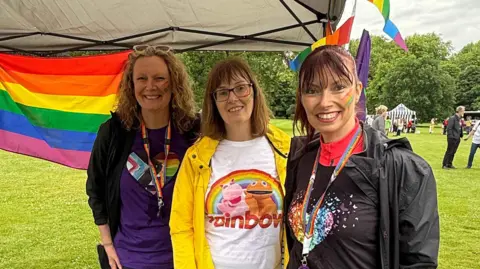 Three people are standing next to each other under a grey gazebo. They are all looking at the camera smiling, and have a rainbow painted on their left cheek. Behind them is a field with people standing on it. 