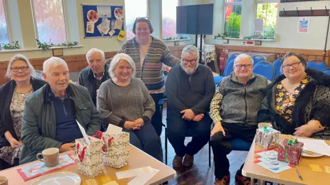 BBC Four women and four men sit in a church hall. One woman stands behind them. They are all wearing winter clothes. There are two tables in front of them with Christmas gift bags, napkins, plates, leaflets and cards on them. Behind them are blue chairs, a television, a noticeboard and six windows. The top half of the walls are cream, the bottom half is covered by wooden panelling.