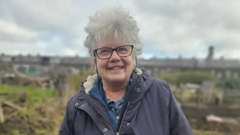 Aisha Iqbal/BBC Tessa Wainwright, one of those involved in the Bradford Refugee and Asylum Seekers Allotment Project, stands smiling in an allotment plot.