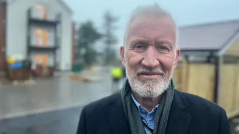 A white man with short white hair and a white beard looks at the camera. He s wearing a dark green scarf and a blue shirt and black coat. Behind him is a block of flats.