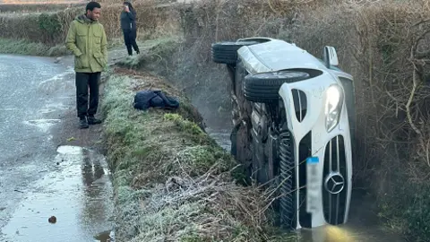 A white Mercedes car overturned on its side in a river stream. A man is seen on the edge of the road staring at the car.