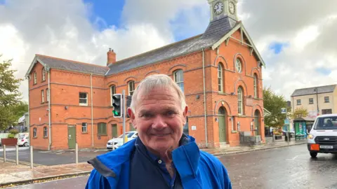 A man wearing a bright blue waterproof jacket and navy blue top is looking into the camera. He has white hair and is smiling. Behind him is a large red brick building with a clock tower. You can also see a grey paved road with a white vehicle travelling toward the camera. 