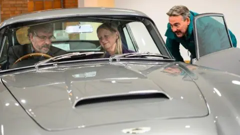 Dominic Fraser/Aston Martin Works John and Susan sit in the silver birch grey car looking at the dashboard as an Aston Martin employee smiles at them from outside the car