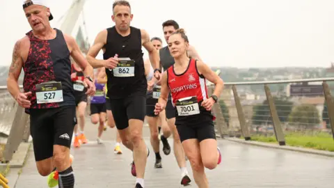 Judith Storm running during the race. She is wearing a red top and black shorts. Running across a bridge.