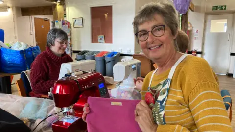 A indoor workspace that appears to be a sewing or crafting room. A woman wearing a mustard-yellow, long‑sleeved, striped sweater and an apron is holding up a pink fabric item, possibly a handmade pouch or bag. The fabric piece has a small rectangular label attached near the top. Another woman is seated at a table, operating a white sewing machine. Next to that machine, there is a red sewing machine, and various pieces of fabric and supplies are scattered across the table.