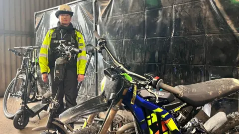 A female police officer dressed in a black uniform, yellow hi-vis vest and white hat stands in a warehouse surrounded by electric bikes and scooters. Behind her is a black screen. The bike closest to the camera resembles a scrambler motorcycle, with a large saddle and colourful cowling.