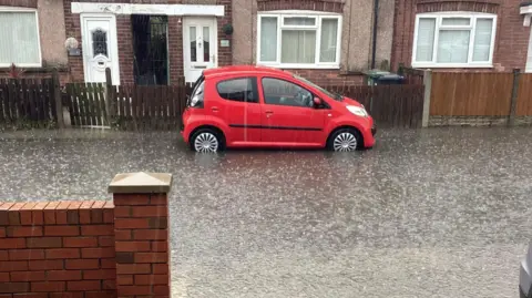 LDRS A bright red car is parked on a street with rain flooding the road.