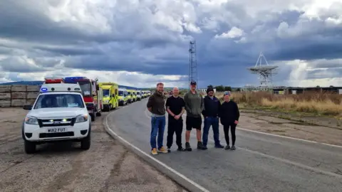 West Mercia Police Five people stood on a road beside a row of parked emergency vehicles, with a large satellite dish on the horizon