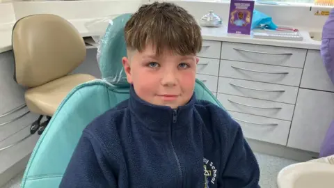 Sitting in a dental chair wearing a navy blue zip up jumper, Kody looks up at the camera. Several drawers and another chair can be seen behind him.