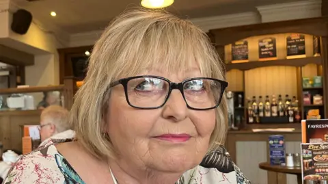 John Devine/BBC A woman facing towards the camera while looking to her right. She is sitting in a pub and people are sat at tables behind her.