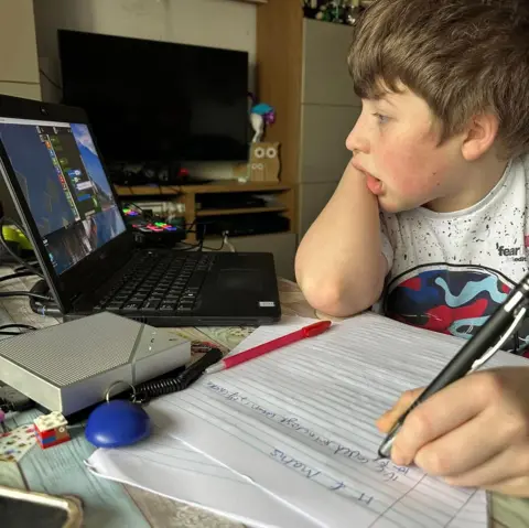 Necessary support group A young boy with brown hair wearing a white t shirt. he is sat at a desk with a pen in his hand and papers in front og him, looking at a laptop. His head is in his hand and he is looking at a black laptop with a fed-up expression on his face. 