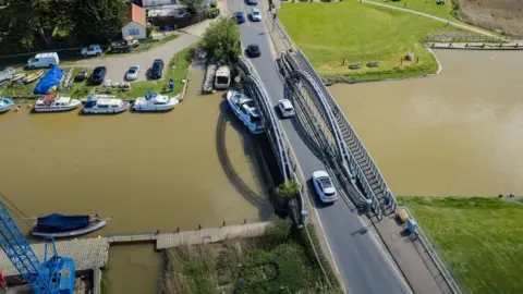 Oliv3r Drone Photography A drone image of a white cruiser wedged under a metal bridge over a river, with a marina in the distance.