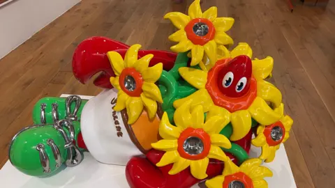 PA Media A sculpture of a cartoon-like lobster head and claws emerging from between a bunch of yellow and orange flowers. The lobster's body is a white and orange flowerpot, with green shoes at the bottom tied with silver laces.