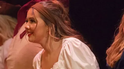 Supplied A close-up of Ella Walden taking part in a drama or dance production. She is sideways on smiling, wearing a white smocked top, with cloth band holding back her long mid brown hair. Behind her can just be seen other people taking part in the same event. 