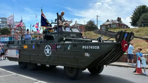 A large military tank is driving down a street. The sun is shining, there are various flags being flown in the background and two children at the front of the tank are standing and admiring it as it drives past.