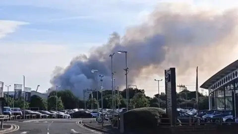 A large plume of smoke can be seen over a treeline, behind a retail park