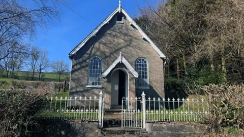 Halls A small chapel with a porch and windows is behind a white metal fence with countryside and trees behind it.