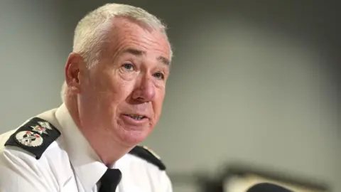 PA Media Jon Boutcher is wearing a white shirt and black tie. He has black patches on his shoulders with three silver badges. He has short grey hair. 