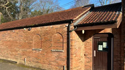 A long brick building, with a wooden door at the end. Behind there are trees peeking out above it.