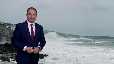 BBC Weather's Stav Danaos standing in front of a picture of the sea