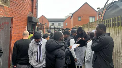 A group of worshippers holding pens and pieces of paper as they gather outside a red-brick building on an overcast day. 