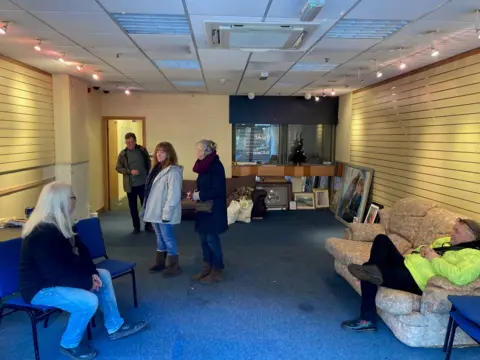 Five people standing and sitting inside an empty shop interior with a blue carpet and yellow walls.  