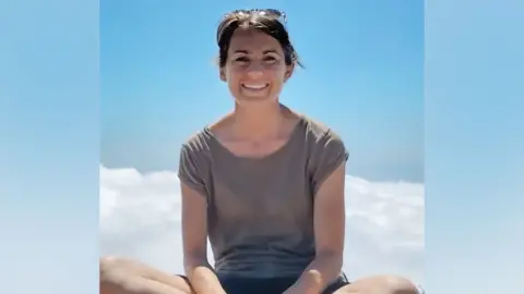 A person outdoors against a clear blue sky. The person is facing the camera and wearing a grey short-sleeved top, with sunglasses resting on their head. Bright daylight creates a soft glow around the figure, and a small area of white cloud is visible in the lower left corner of the image.