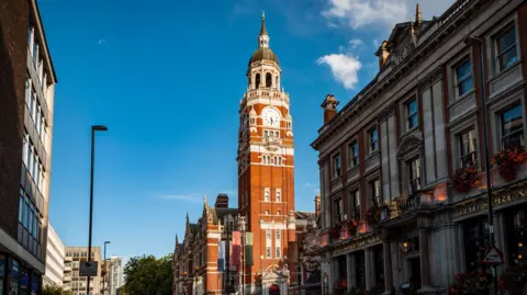 A red Victorian brick clocktower rises above buildings in Croydon town centre. It is a sunny with a blue sky. 