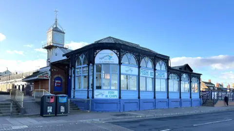 The Mermaid fish and chip cafe - a Victorian-era wood and ironwork building with large ornate windows. It has been painted blue with black ironwork. A clock tower and railway station can be seen in the distance.