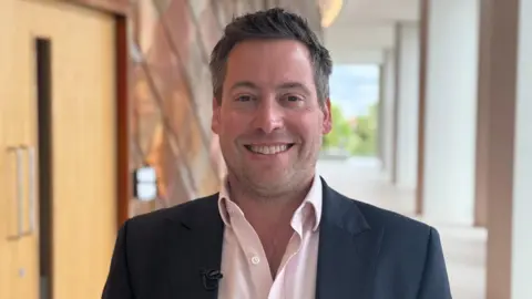 Dr Dave Ellis is wearing a dark blue suit and open-necked pink shirt. He is standing in a corridor at the Ray Dolby Centre smiling at the camera. There is a lecture hall lined with copper plates on one side and large windows on the other.