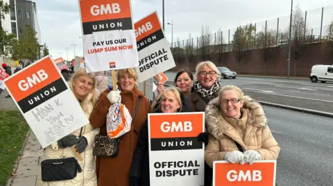 Members of the GMB union and hospital staff stand on the picket line outside Royal Liverpool University Hospital holding placards saying 'Pay us our lump sum now' and 'Official dispute'.