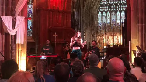 BBC A woman with long dark hair in a cropped top sings into a microphone. The backs of people's heads can be seen in the foreground, and a large stained glass window behind