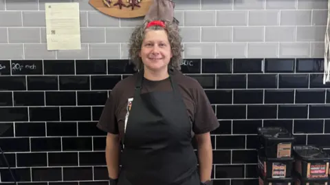 Lilith is standing against black tiles in her shop on the high street. She has brown curly hair pulled back with a red band and a black apron on over a brown T'shirt.
