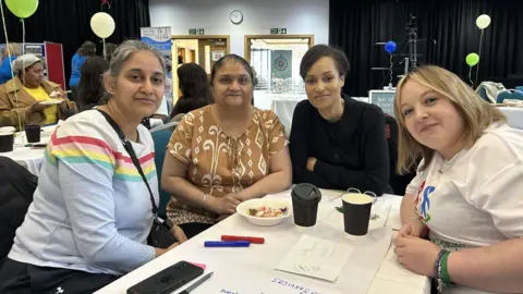 Four women seated around a table in a large room with balloons on tables. From left to right there is a woman with grey hair and wearing a pale blue top with stripes on it and a handbag crossbody. Next to her is a woman with dark hair and a patterned brown top. To her right is a woman with short dark hair and a dark long sleeved top. And next to her is a woman with blonde hair and a white top. They are all looking at the camera and smiling. 