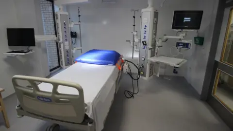 University of Bath An empty medical room, with white walls and a grey floor. An inflated blue medical pillow, connected to tubes, is displayed on top of a medical bed that has only a white sheet.