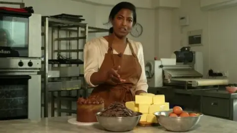 Gaya Vara Gaya Vara look at the camera wearing an apron and standing in a kitchen with various bakery ingredients in front of her including a pile of butter, eggs and chocolate.