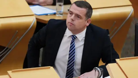 PA Media Douglas Ross sitting in the Scottish Parliament, with his left arm resting on a desk. He is wearing a black suit, white shirt and blue and black striped tie.