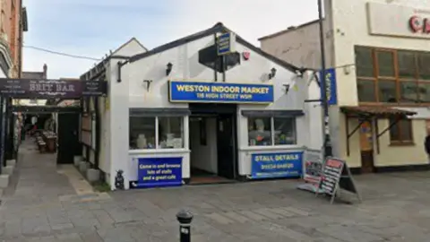 Google A Google Street View image shows the front of Weston Indoor Market. A single storey building, a blue sign above the door reads "Weston Indoor Market. 116 High Street WSM". 
