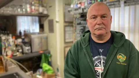 Craig Armstrong wearing a green top and blue T-shirt. A bar area is blurred in the background.