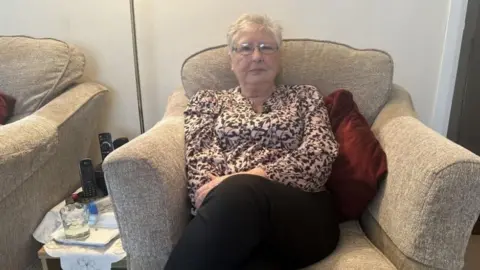 An elderly woman with short grey hair, wearing spectacles and a brown and cream patterned top. Her expression is serious and she is sitting in a large light brown cushioned arm chair.