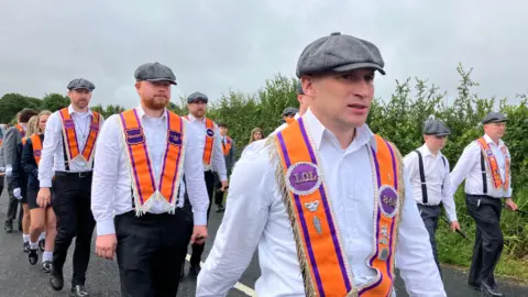 Men marching in white shirts and with orange sashes around their shoulders and grey flat caps