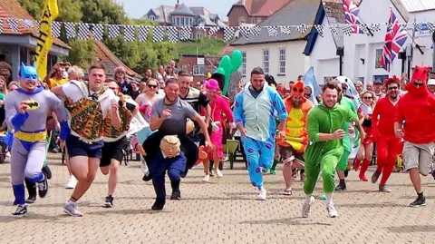 A line of competitors in the Watchet wheelbarrow races smile and shout as they sprint towards the camera. Many of them are in fancy dress